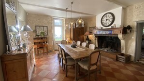A dining area with a checkered floor, wooden table, chairs, and pendant lights. A fireplace with a clock above it is visible.