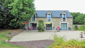 Un bâtiment en pierre avec une allée gravillonnée, des sièges à l’extérieur et un grand arbre.
