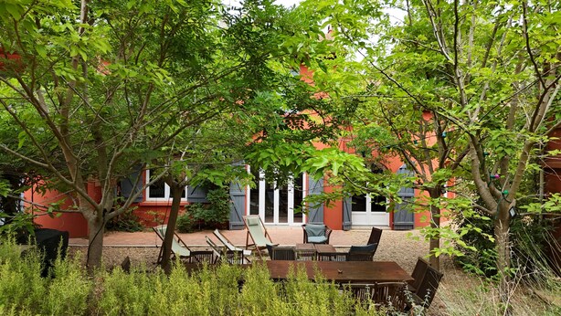 Un espace extérieur aménagé avec des tables et des chaises, entouré d’arbres et bordé d’un bâtiment rouge doté de fenêtres.