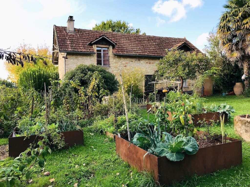 Une maison en pierre avec un toit en tuiles, entourée d’un jardin bien entretenu, agrémenté de parterres surélevés et de diverses plantes.