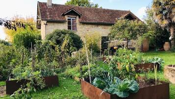 Une maison en pierre avec un toit en tuiles, entourée d’un jardin bien entretenu, agrémenté de parterres surélevés et de diverses plantes.