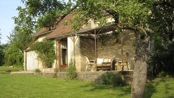 Une maison de campagne en pierre, avec un toit en tuiles, une véranda en bois et un petit jardin agrémenté d’un banc et d’une table.