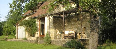 A rustic stone house with a tiled roof, a wooden porch, and a small garden area with a bench and a table.