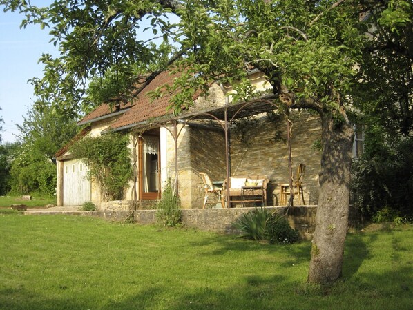 A rustic stone house with a tiled roof, a wooden porch, and a small garden area with a bench and a table.