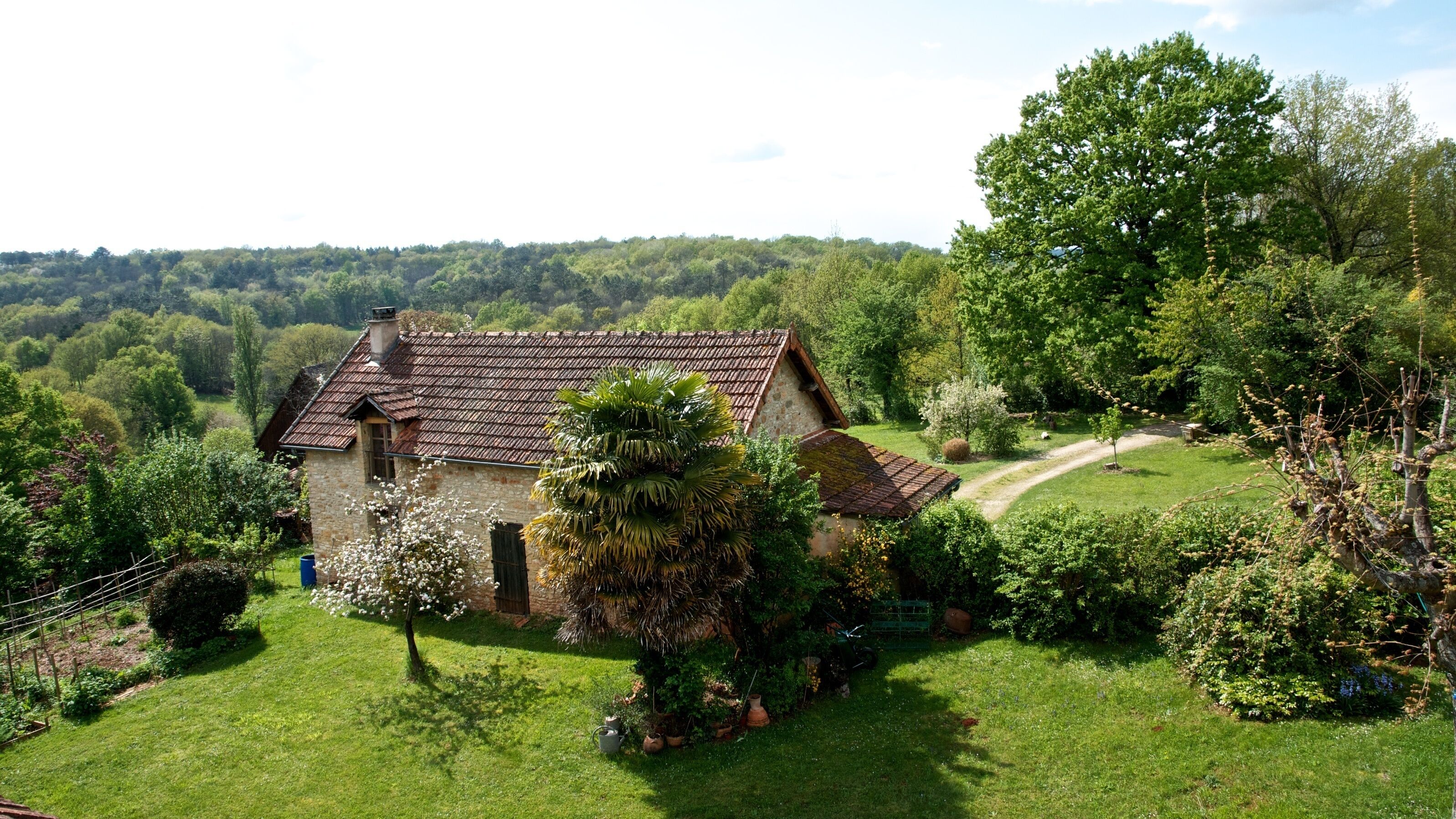 Une maison en pierre, avec un toit de tuiles, entourée d’une végétation luxuriante et accessible par un chemin dégagé.