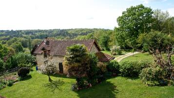 Une maison en pierre, avec un toit de tuiles, entourée d’une végétation luxuriante et accessible par un chemin dégagé.