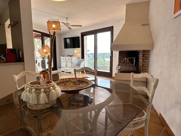 Un coin repas avec une table ronde en verre, des chaises blanches, un panier en osier et un ventilateur de plafond.