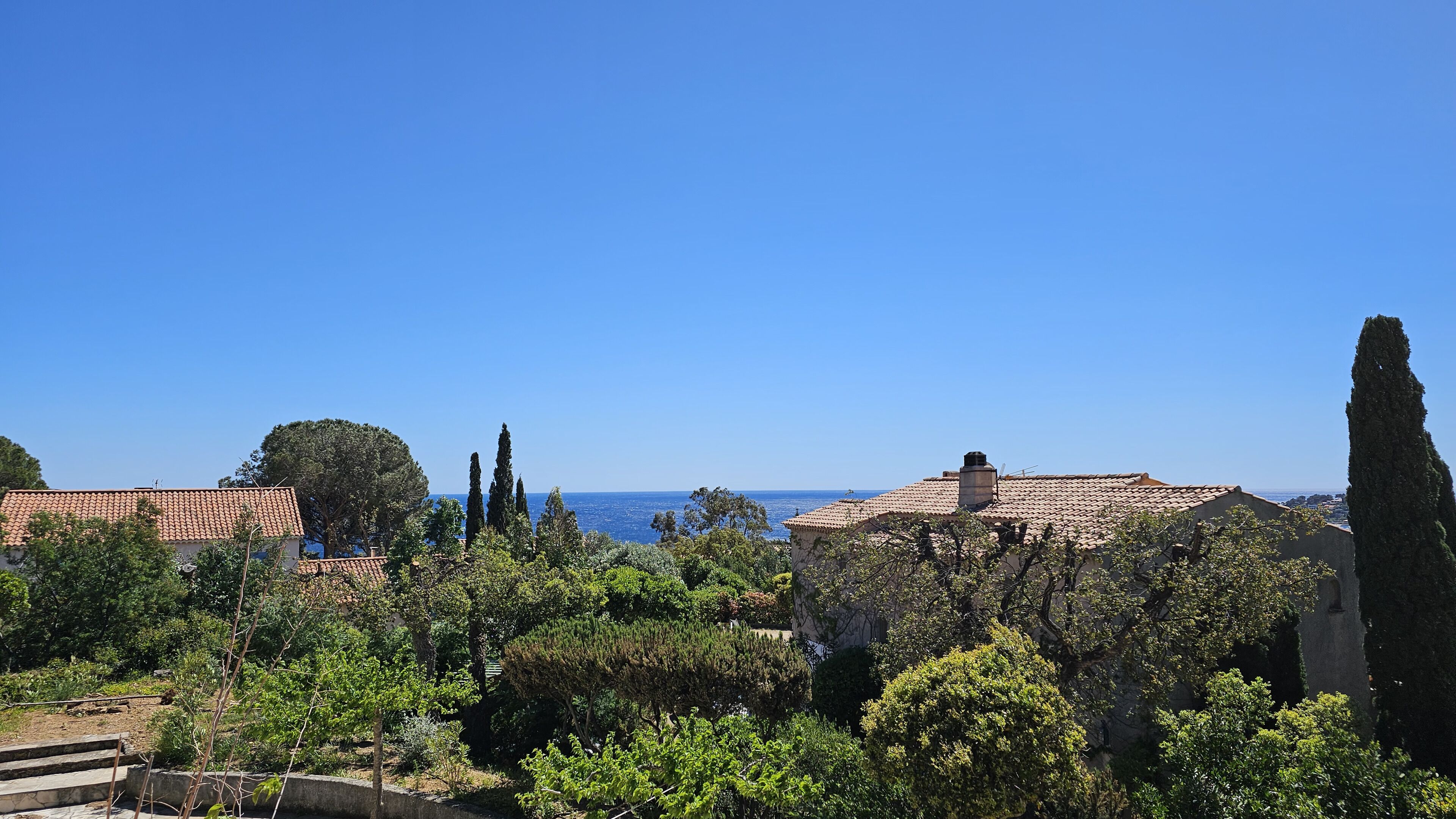 Una zona residencial costera con casas de tejas de color terracota, abundante vegetación y un cielo azul despejado.
