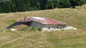 A rustic, single-story stone building with a slanted roof, surrounded by a grassy field and a forested area in the background. - Lelex Monts-Jura to rent studio facing south and resort center  (Lélex)