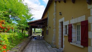 A covered outdoor area with stone flooring, a yellow building with red shutters, and a stone wall with greenery. - Beautiful house of 315 m² in a 4000 m² park with swimming pool 3 minutes from Périgueux (Champcevinel)