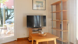 A living room with a TV on a stand, a wooden table, a bookshelf, and a window with a view of a red building and trees. - House with garden beach Scudo (Ajaccio)