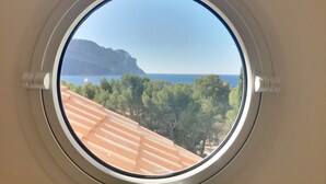 Une fenêtre ronde offrant une vue sur un paysage côtier, avec des arbres et une étendue d’eau.
