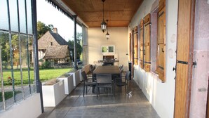 Un porche cubierto con una mesa y sillas, un colgante de luz y vista a una casa con techo de paja.