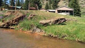 A rustic cabin with a porch, surrounded by a grassy hillside and a small stream. - ~ Cozy Creek Side Cabin ~ Nature ~ Peaceful ~ Relax ~Family!  (Hill City)