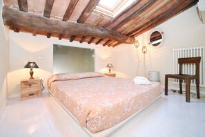 Quarto com cama grande, vigas de madeira, claraboia, espelho, radiador e abajures.