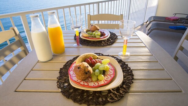 Een tafel met ontbijtproducten, een fles sinaasappelsap en een glas water op een balkon met uitzicht op zee.