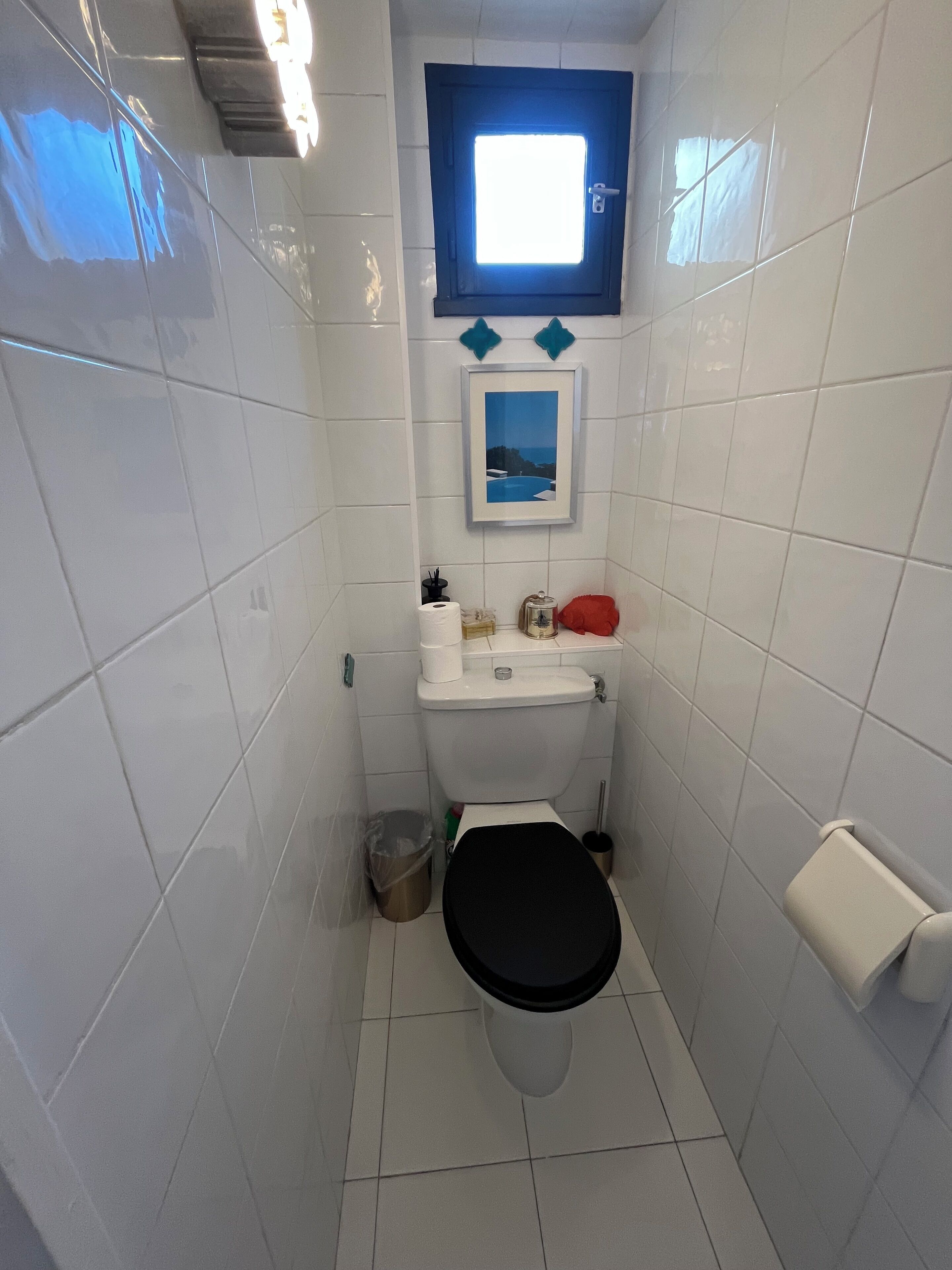 Et lille, kompakt badeværelse med hvide fliser, et sort toilet, en hvid vask og et billede i ramme over vasken.