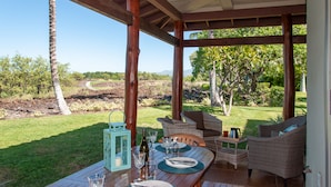 Un área de comedor al aire libre con una mesa lista para dos personas, rodeada de muebles de mimbre, con vista a un jardín ajardinado y colinas lejanas.
