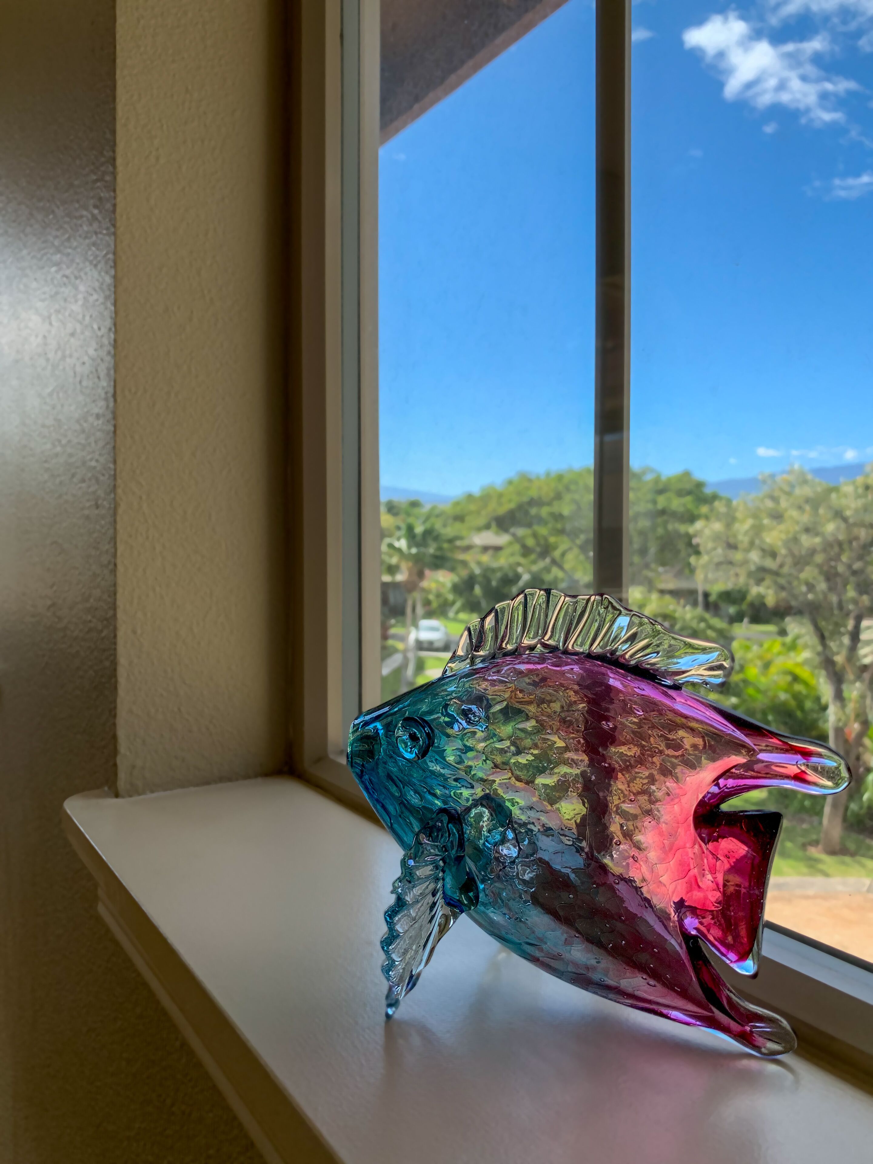 Un pez de vidrio de colores sobre un alféizar con vista a árboles y un cielo despejado.