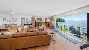 A modern living room with a large leather sofa, a dining area, and a kitchen. A large window offers a view of a lake and mountains.