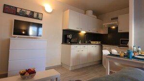 Cocina compacta con armarios blancos, microondas y cafetera. Hay una mesa de comedor con algunos objetos encima, y una mesita con un cuenco de fruta.