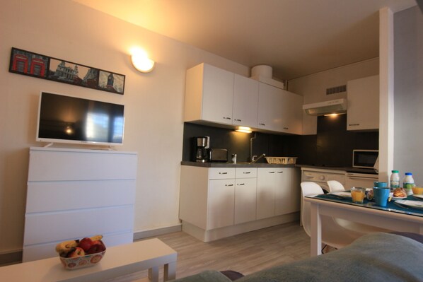 Cocina compacta con armarios blancos, microondas y cafetera. Hay una mesa de comedor con algunos objetos encima, y una mesita con un cuenco de fruta.