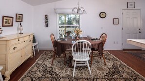 Uma sala de jantar com mesa marrom, cadeiras brancas, um lustre e uma janela grande.