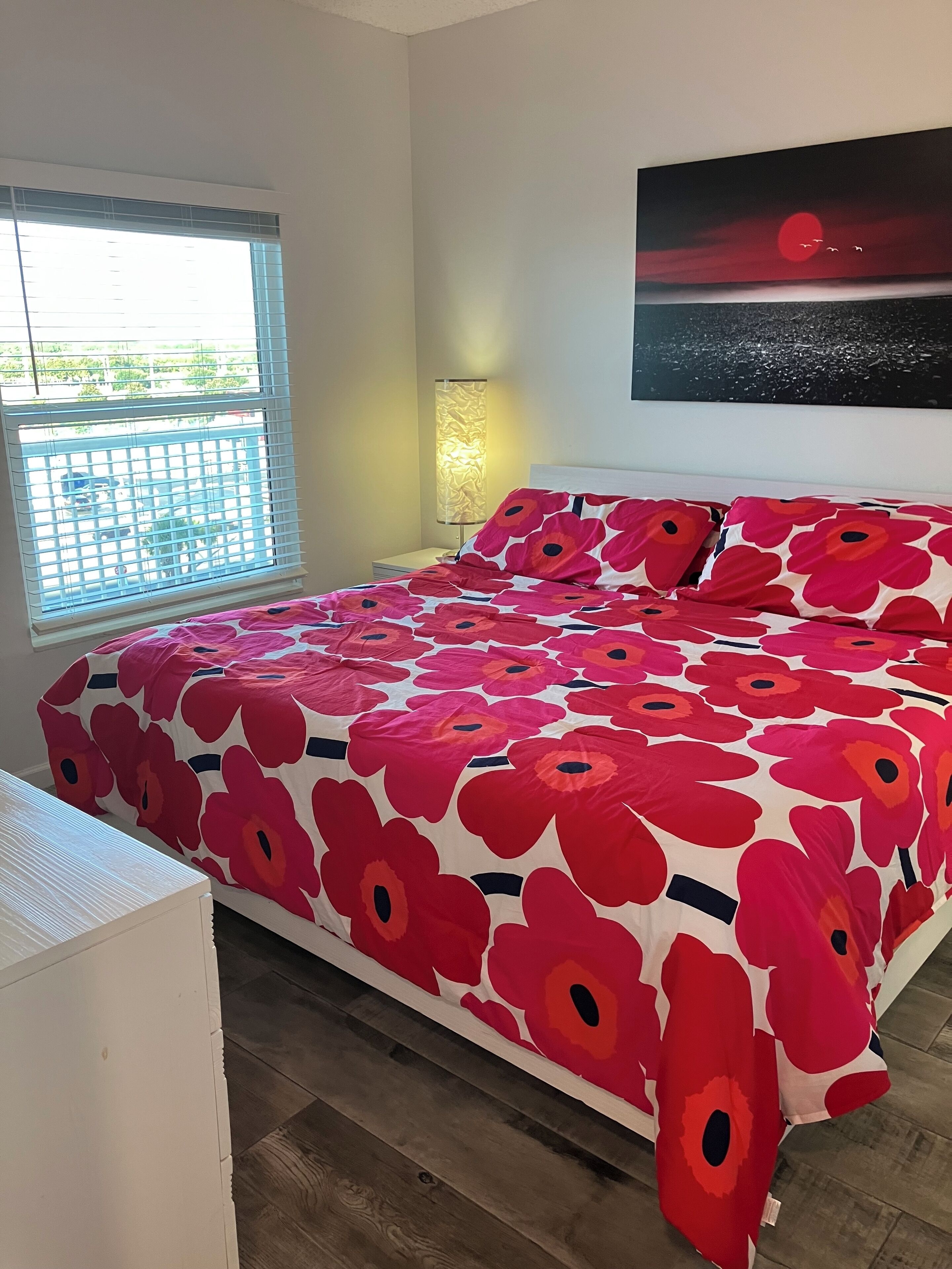 A bedroom with a large bed, a nightstand, a lamp, a window with blinds, and a painting on the wall.