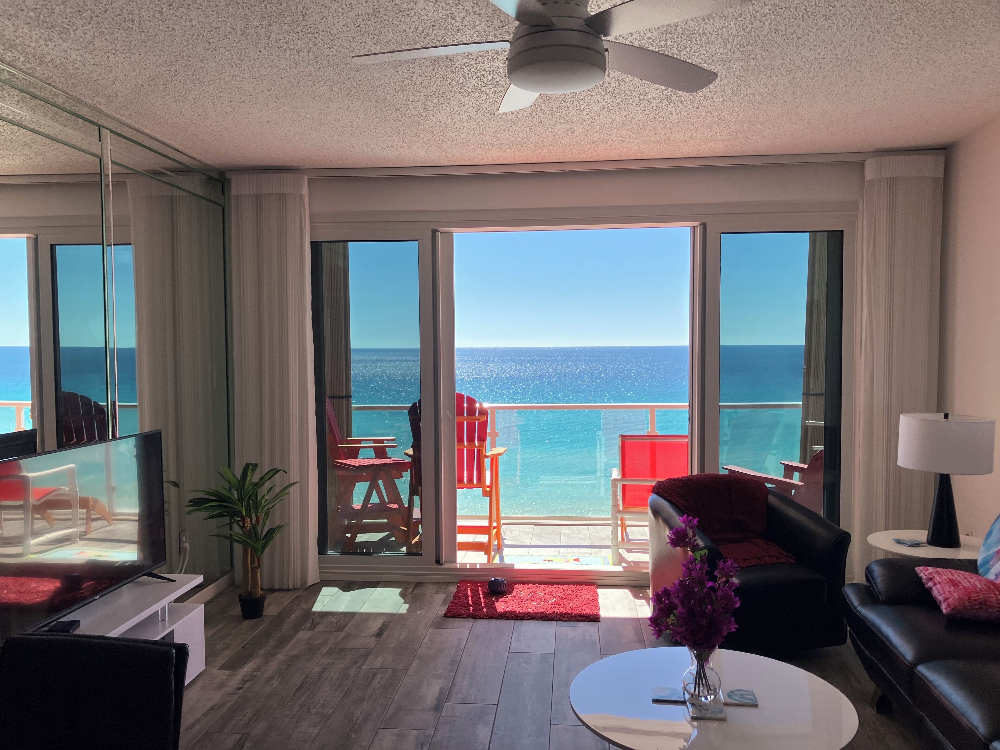A modern living room with a large TV, a ceiling fan, and a view of the ocean through large sliding doors.
