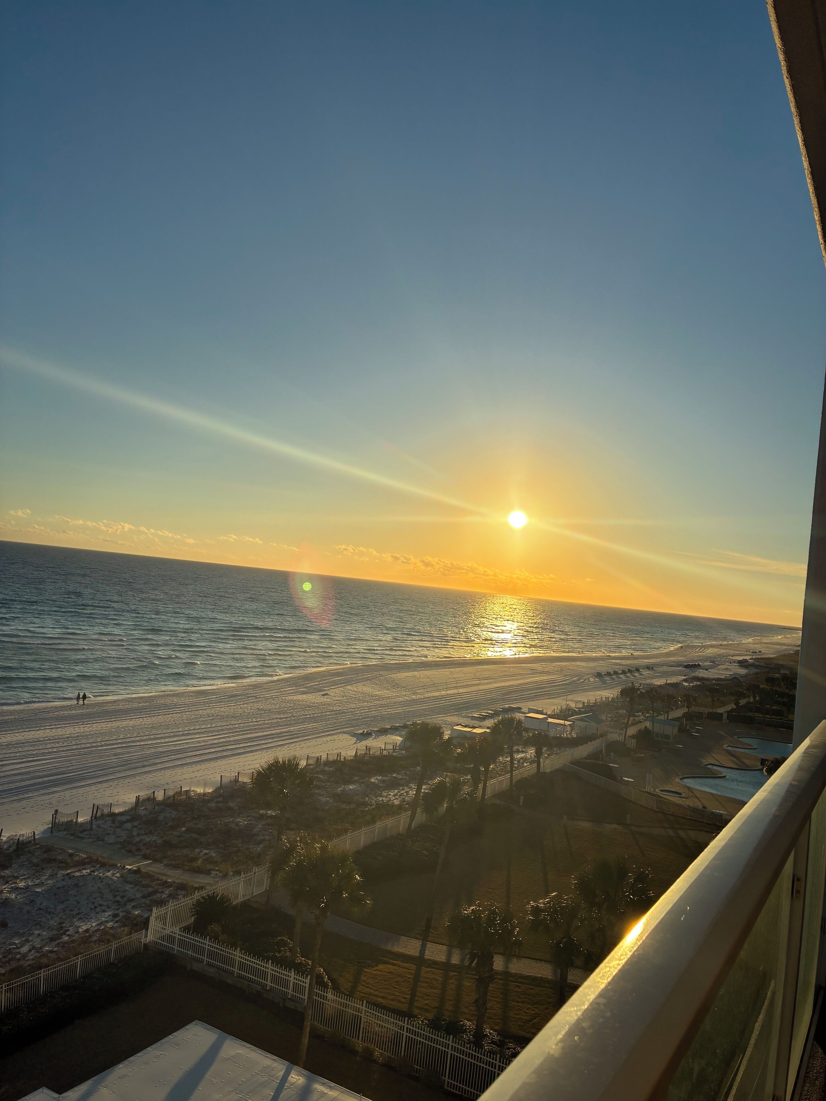 A sunset view from a balcony overlooking a beach with a fence and palm trees.