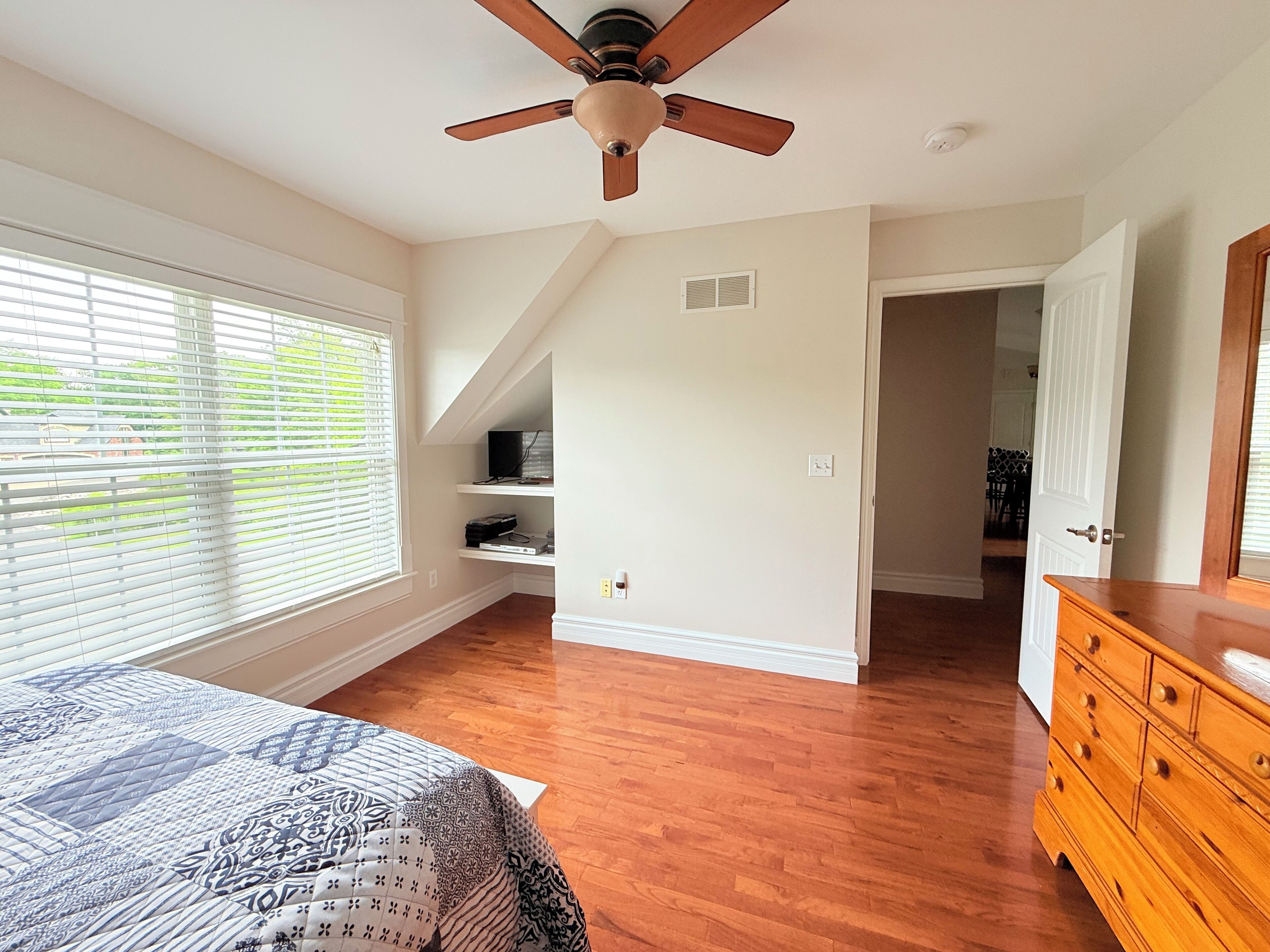 A bedroom with a bed, wooden dresser, ceiling fan, and a window with blinds.