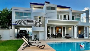A modern two-story house with a swimming pool, a covered garage, and a patio with outdoor furniture. - OCEAN VIEW VILLA WITH PRIVATE POOL ,STEPS FROM SECLUDED BEACH, GATED COMMUNITY! (Yabucoa)