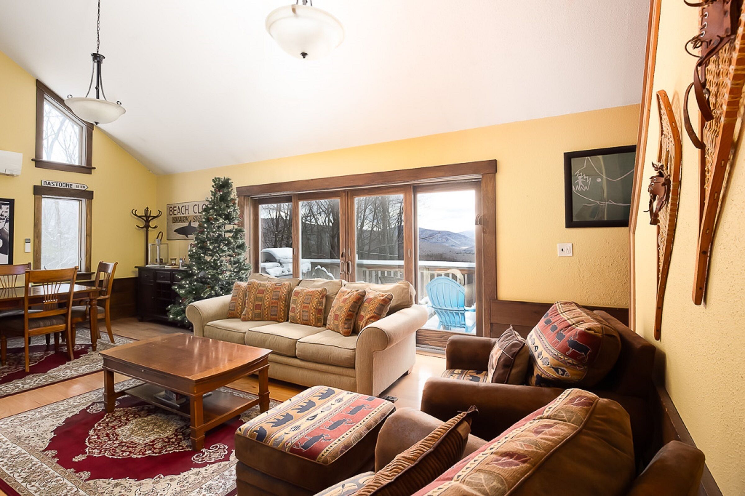 Un salon avec un canapé beige, une table basse et un sapin de Noël. Il y a une fenêtre donnant sur les montagnes et une porte portant l’inscription « Bastogne ».