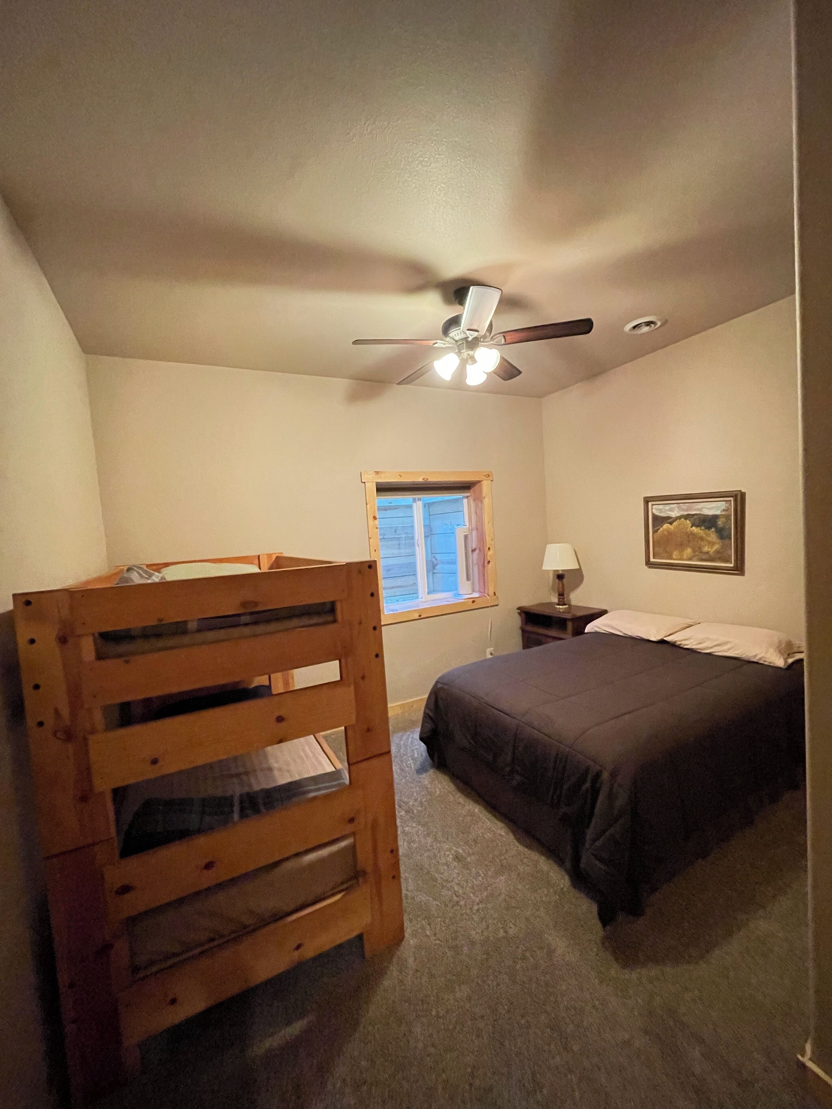 A bedroom with a bunk bed, a single bed, a ceiling fan, a window, a nightstand, a lamp, and a framed picture on the wall.