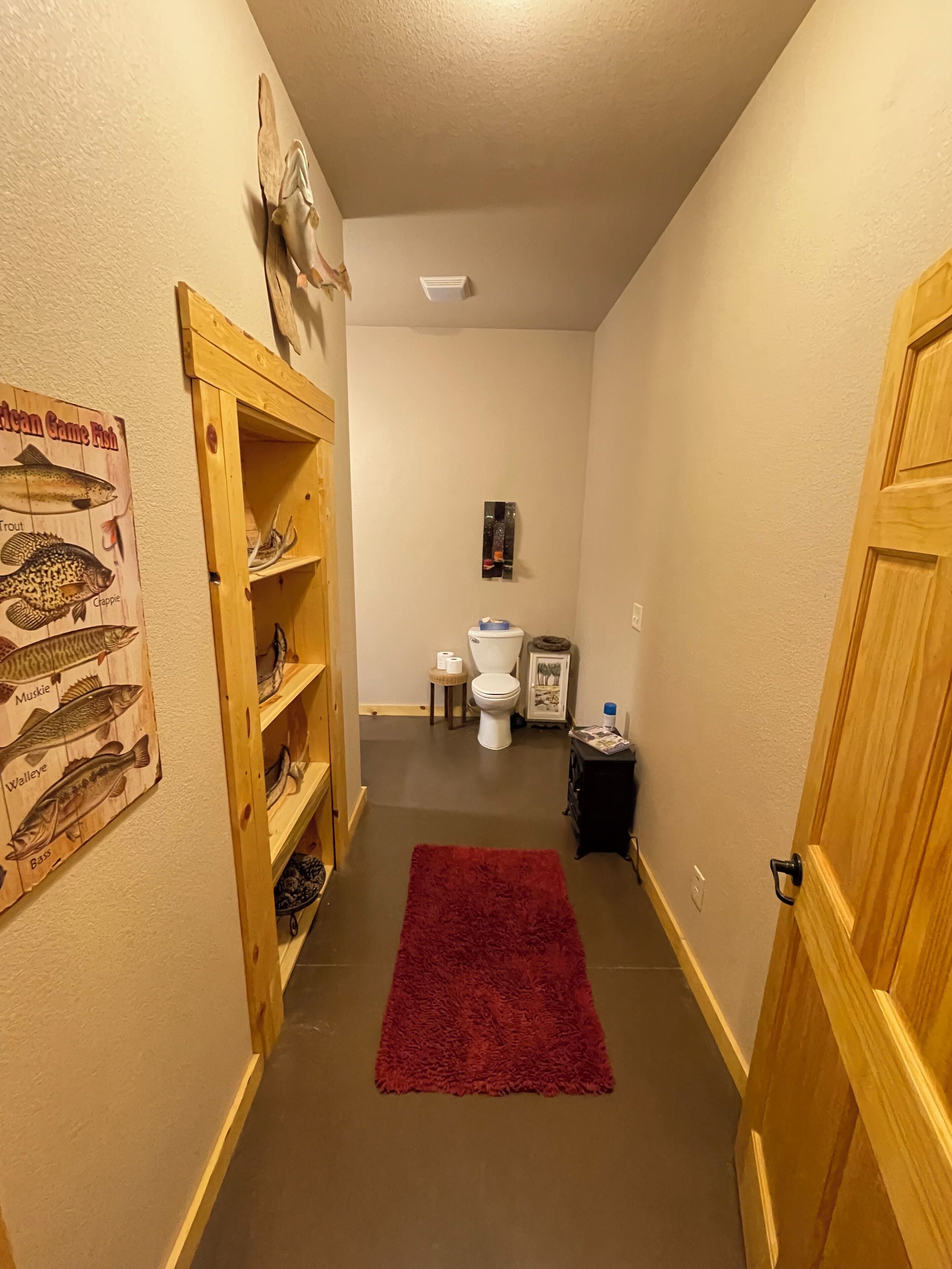 Un pasillo estrecho con una puerta de madera a la derecha, una alfombra roja, un estante de madera con un cartel de un pez, un inodoro y una mesita con un marco de fotos y una botella.