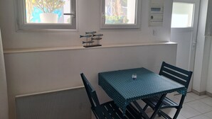 Un pequeño comedor con una mesa y sillas, dos ventanas con cortinas, un radiador y una lámpara fijada a la pared.