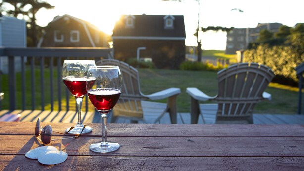 A wooden deck with two wine glasses and a pair of sunglasses on a table, surrounded by outdoor chairs and a house in the background. - Charming Cannon Beach Cottage- Perfect Family Getaway near the beach! (Cannon Beach)