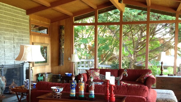 Un salon chaleureux avec une cheminée, des poutres en bois, une grande fenêtre donnant sur les arbres, un canapé rouge et une table basse en bois agrémentée d’objets décoratifs.