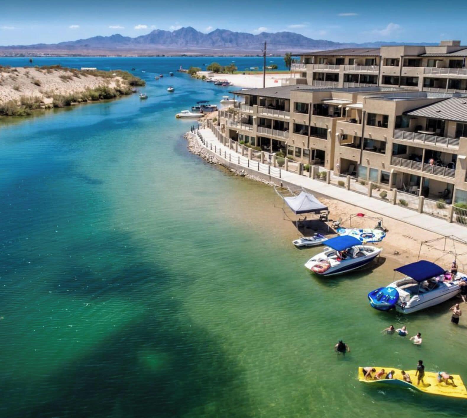 Un hotel frente al mar con un puerto deportivo, aguas azules cristalinas y montañas al fondo.