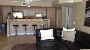A modern living room with a black leather sofa, a kitchen area with a central island, and a ceiling fan.