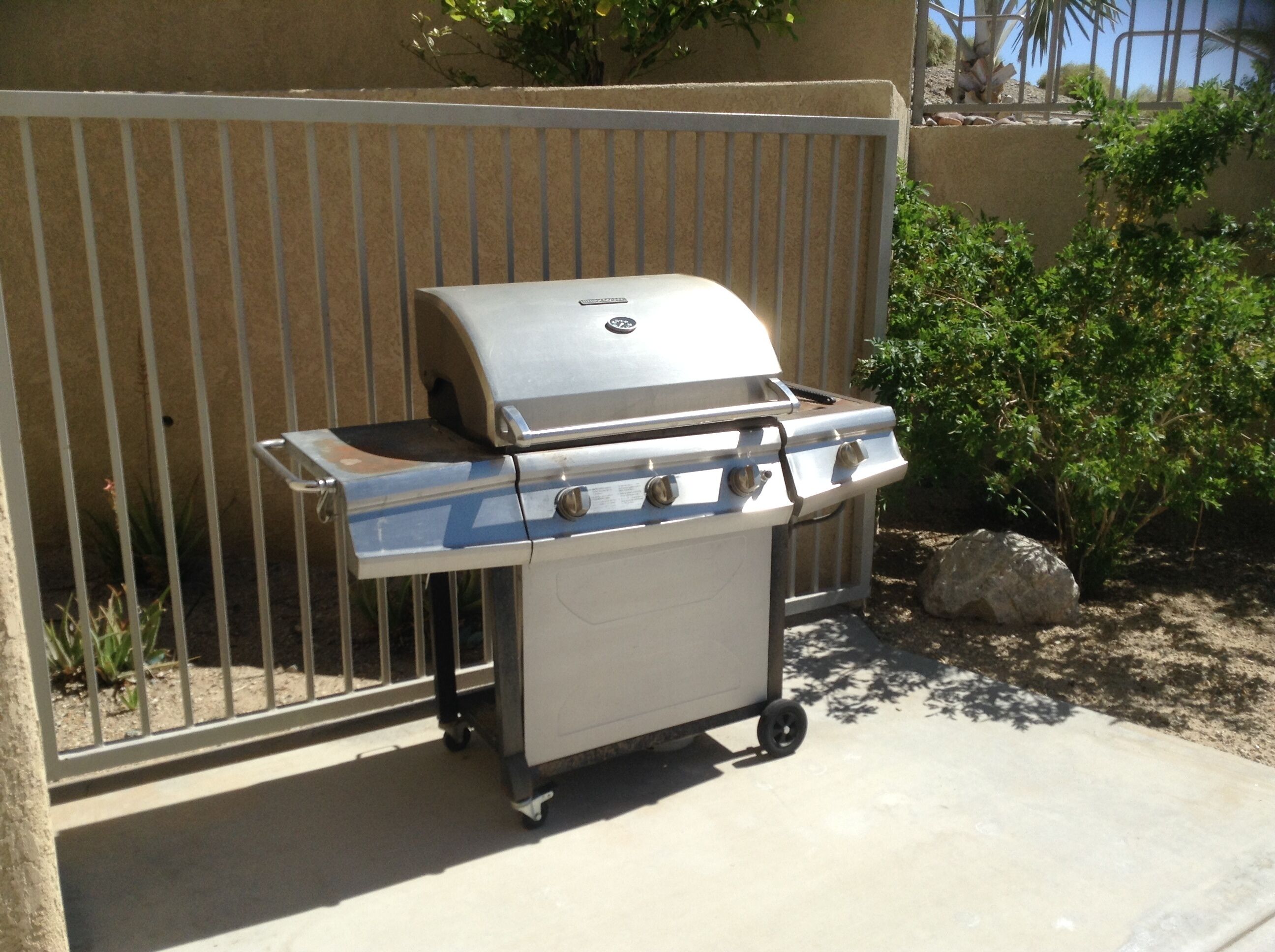 A portable gas grill with four burners, a side shelf, and a lid, placed on a concrete surface near a metal fence.