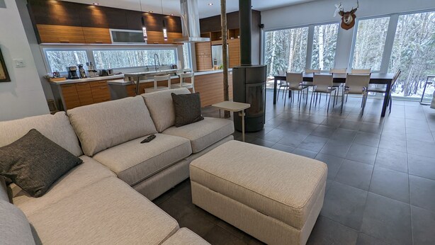 A modern living room with a large beige sectional sofa, a small round table, and a dining area with chairs. - Treehouse, 3 bedrooms (4th bedroom, sleeps 7+), EV terminals. (Petite-Rivière-St-Francois)