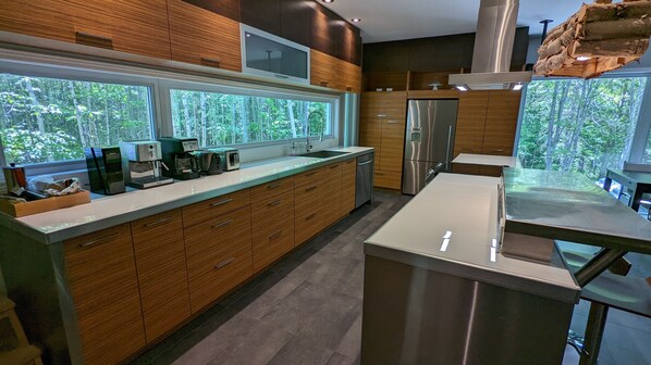 A modern kitchen with wooden cabinets, a stainless steel refrigerator, and a large window overlooking greenery. - Treehouse, 3 bedrooms (4th bedroom, sleeps 7+), EV terminals. (Petite-Rivière-St-Francois)