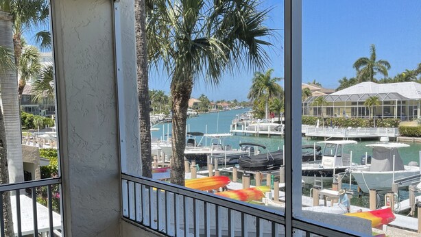A balcony with a ceiling fan, a white chair, and a view of a marina with boats and palm trees.