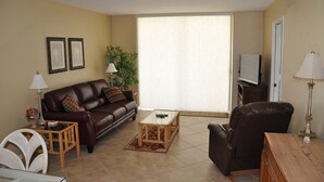 A living room with a brown leather sofa, a glass coffee table, a wooden side table, a brown leather chair, a TV, and a large window with blinds.