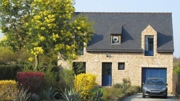 Une maison en pierre avec une voiture garée devant, entourée de différentes plantes et arbres.