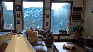 A cozy living room with a leather sofa, a table, and a lamp. There are framed pictures on the wall and a view of a forest through large windows. - Spearfish canyon vacation home 15 minutes from Terry Peak Ski Area & Deadwood. (Lead)