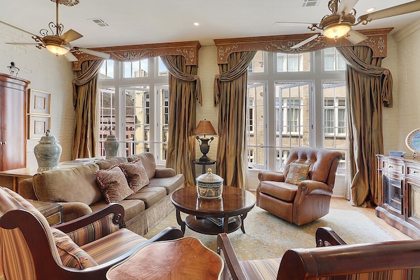 A spacious living room with a sofa, armchairs, a coffee table, and a fireplace. Large windows with curtains provide natural light and views of the city.