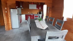 Interior de una cabaña de madera con una mesa de comedor, sillas, una zona de cocina y una ventana con cortinas.