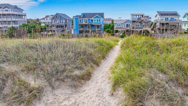 Una zona residenziale costiera con diverse case a due piani, un sentiero sabbioso che si snoda tra le dune e un cielo azzurro limpido.